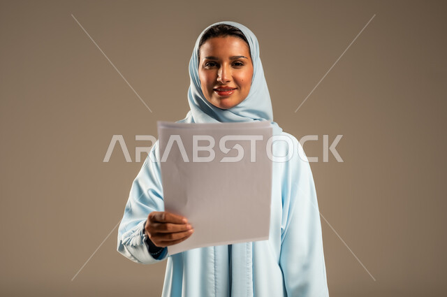 مفهوم إداة الأعمال والمشاريع، تعابير السعادة والسرور، بورتريه مقرب لامراة عربية خليجية سعودية محجبة ترتدي عباءة ملونة تحمل بيدها مجموعه من الاوراق تنظر الى الكاميرا بايماءات الثقة بالنفس، الأناقة والاهتمام بالمظهر الخارجي، خلفية رمادية