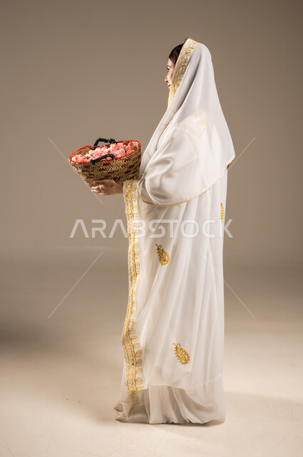 يوم بدينا 1727 م، المناسبات والأعياد الوطنية، ذكرى تأسيس الدولة السعودية الاولى 22 فبراير، بورتريه من الجانب لشابة عربية خليجية سعودية ترتدي العباءة البيضاء تحمل بيدها سلة من القش مليئة بالورد الجوري الطبيعي، صورة الجسم بالطول الكامل، خلفية بيج