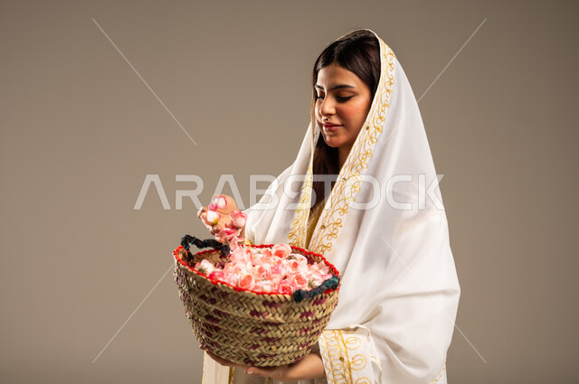 Anniversary of the founding of the first Saudi state, February 22, close-up portrait of a young Saudi Arabian Gulf woman wearing a white abaya, holding a straw basket filled with natural roses, the day we began 1727 AD, the concept of love of the homeland and belonging to it, beige background