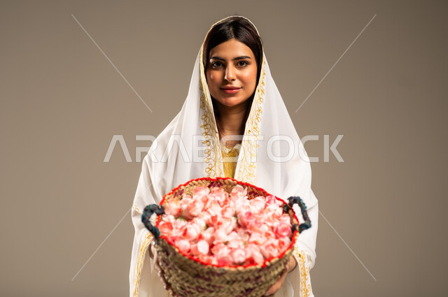 ذكرى تأسيس الدولة السعودية الاولى 22 فبراير، بورتريه مقرب لشابة عربية خليجية سعودية ترتدي العباءة البيضاء تنظر الى الكاميرا تحمل بيدها سلة من القش مليئة بالورد الجوري الطبيعي، يوم بدينا 1727 م، مفهوم حب الوطن والانتماء اليه، خلفية بيج