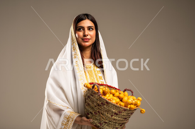 يوم بدينا 1727 م، منتجات ومحاصيل محلية، ذكرى تأسيس الدولة السعودية الاولى 22 فبراير، المناسبات والأعياد الوطنية، بورتريه مقرب لشابة عربية خليجية سعودية ترتدي العباءة البيضاء تحمل بيدها سلة من القش مليئة بحبات البلح، خلفية بيج