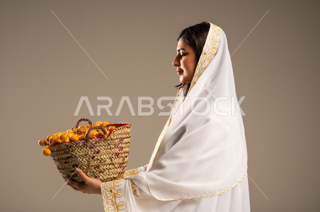 The Day We Began 1727 AD, Local Products and Crops, Anniversary of the Founding of the First Saudi State February 22, National Events and Holidays, Close-up portrait from the side of a young Saudi Arabian Gulf woman wearing a white abaya holding a straw basket full of dates, beige background