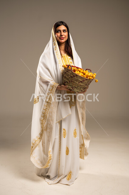 منتجات ومحاصيل محلية، ذكرى تأسيس الدولة السعودية الاولى 22 فبراير، المناسبات والأعياد الوطنية، بورتريه لشابة عربية خليجية سعودية ترتدي العباءة البيضاء تحمل بيدها سلة من القش مليئة بحبات البلح، يوم بدينا 1727 م، صورة الجسم بالطول الكامل, خلفية بيج