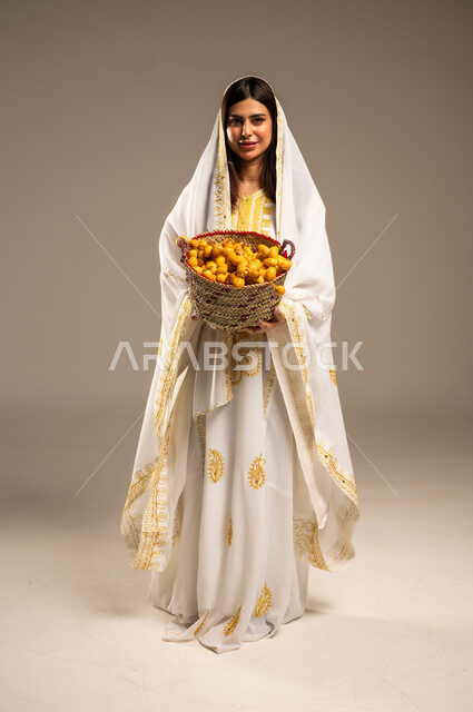 Anniversary of the founding of the first Saudi state February 22, local products and crops, national occasions and holidays, portrait of a young Saudi Arabian Gulf woman wearing a white abaya looking at the camera holding a straw basket full of dates, the day we started 1727 AD, full body image, beige background