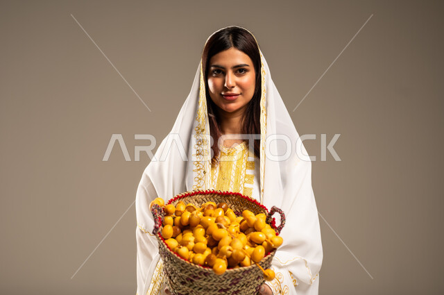 ذكرى تأسيس الدولة السعودية الاولى 22 فبراير، الاحتفال بالمناسبات والأعياد الوطنية، بورتريه مقرب لشابة عربية خليجية سعودية ترتدي العباءة البيضاء تنظر الى الكاميرا تحمل بيدها سلة من القش مليئة بحبات البلح، يوم بدينا 1727 م، منتجات ومحاصيل محلية، خلفية بيج
