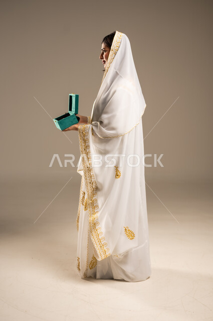 Distinctive aromatic scents, the anniversary of the founding of the first Saudi state, February 22, celebrating national occasions and holidays, portrait of a young Saudi Arabian Gulf woman wearing a white abaya holding a box of oud wood, the day we started 1727 AD, full-length body image, beige background