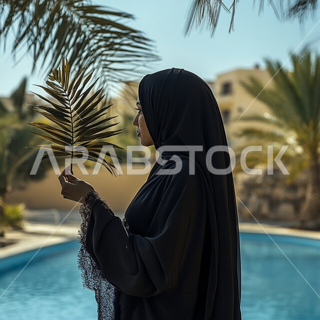أزياء محجبات عصرية أنيقة, الاهتمام بالأناقة و المظهر الخارجي, مفهوم الأنوثة و النعومة, صورة من الجانب لإمرأة عربية خليجية سعودية محجبة ترتدي العباءة السوداء تقف أمام المسبح و تمسك بيدها غصن شجرة