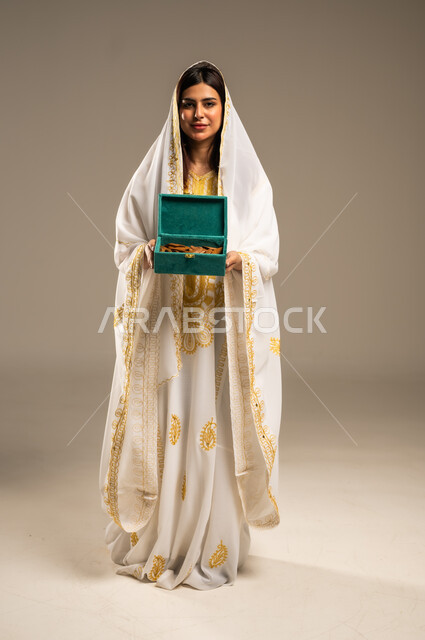روائح عطرية متميزة، ذكرى تأسيس الدولة السعودية الاولى 22 فبراير، الاحتفال بالمناسبات والأعياد الوطنية، بورتريه لشابة عربية خليجية سعودية ترتدي العباءة البيضاء تنظر الى الكاميرا تحمل بيدها صندوق من خشب العود، يوم بدينا 1727 م، صورة الجسم بالطول الكامل، خلفية بيج