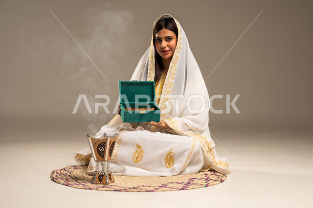 The Day We Began 1727 AD, Distinctive Aromatic Smells, Anniversary of the Founding of the First Saudi State February 22, Portrait of a young Saudi Arabian Gulf woman wearing a white abaya holding a box of oud wood in her hand, sitting on a straw mat in front of the incense burner, looking at the camera with gestures of pleasure, beige background