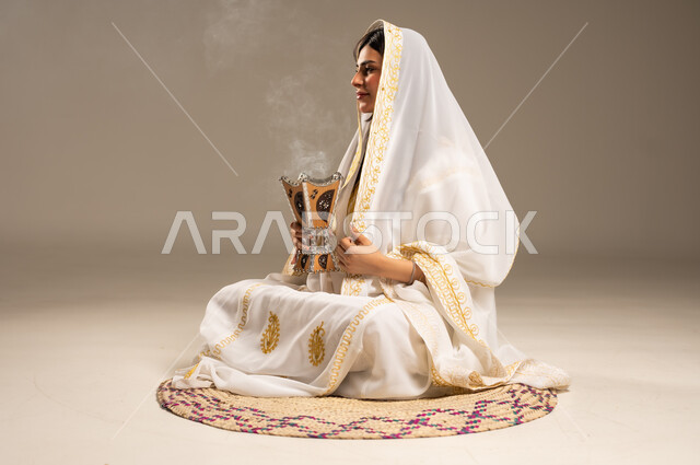 Using agarwood to perfume with incense, the anniversary of the founding of the first Saudi state, February 22, a portrait of a young Saudi Arabian Gulf woman wearing a white abaya sitting on a straw mat holding an incense burner in her hand, the day we began 1727 AD, beige background