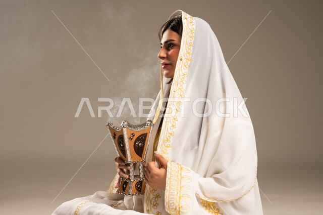 Using agarwood for incense, celebrating national occasions and holidays, the anniversary of the founding of the first Saudi state, February 22, a side portrait of a young Saudi Arabian Gulf woman wearing a white abaya sitting holding an incense burner in her hand, the day we began 1727 AD, the concept of love of the homeland and belonging to it, beige background