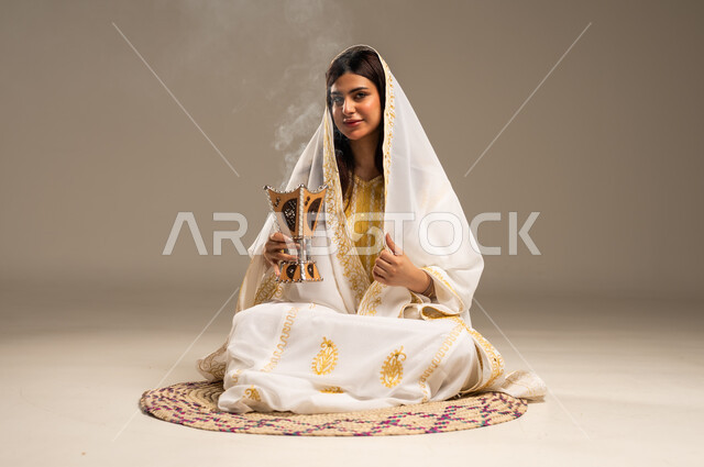 استخدام خشب العود للتطيب بالبخور، ذكرى تأسيس الدولة السعودية الاولى 22 فبراير، بورتريه لشابة عربية خليجية سعودية ترتدي العباءة البيضاء تجلس على حصيرة من القش تحمل بيدها المبخرة تنظر الى الكاميرا بإيماءات السرور، يوم بدينا 1727 م، خلفية بيج
