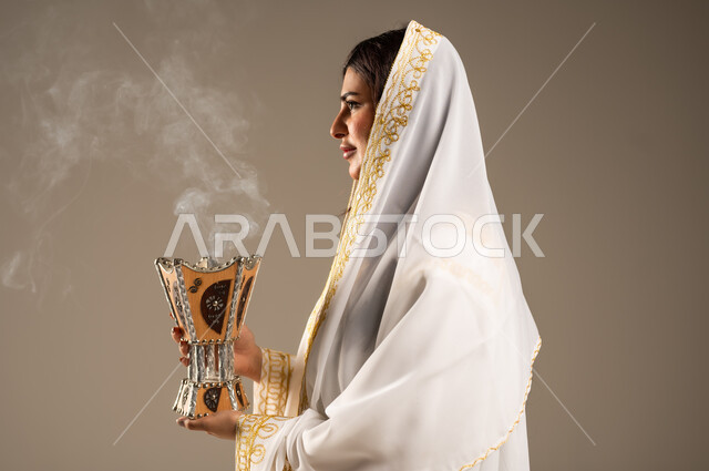 استخدام خشب العود للتطيب بالبخور، الاحتفال بالمناسبات والأعياد الوطنية، ذكرى تأسيس الدولة السعودية الاولى 22 فبراير، بورتريه مقرب من الجانب لشابة عربية خليجية سعودية ترتدي العباءة البيضاء تحمل بيدها المبخرة، يوم بدينا 1727 م، خلفية بيج