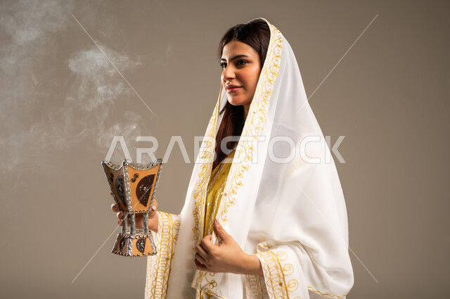 Using agarwood for incense, celebrating national occasions and holidays, the anniversary of the founding of the first Saudi state, February 22, a close-up portrait of a young Saudi Arabian Gulf woman wearing a white abaya holding an incense burner, the day we began 1727 AD, beige background
