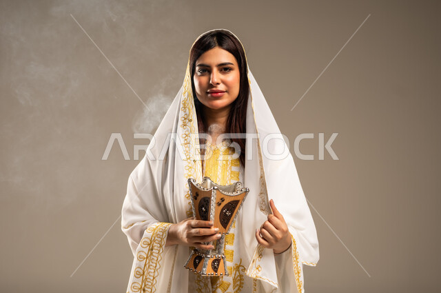 Using agarwood for incense, celebrating national occasions and holidays, the anniversary of the founding of the first Saudi state, February 22, a close-up portrait of a young Saudi Arabian Gulf woman wearing a white abaya looking at the camera holding an incense burner in her hand, the day we began 1727 AD, beige background