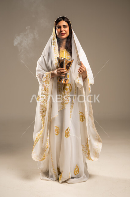 The Day We Began 1727 AD, Using Oud Wood for Incense, Celebrating National Occasions and Holidays, Anniversary of the Founding of the First Saudi State February 22, Portrait of a Young Saudi Arabian Gulf Woman Wearing a White Abaya Looking at the Camera Holding an Incense Burner in Her Hand, Full-Length Body Image, Beige Background
