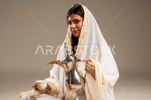 مفهوم الكرم وحسن الاستقبال، يوم بدينا 1727 م، الاحتفال بالمناسبات والأعياد الوطنية، ذكرى تأسيس الدولة السعودية الاولى 22 فبراير، بورتريه مقرب لشابة عربية خليجية سعودية ترتدي العباءة البيضاء تقوم بصب القهوة من الدلة النحاسية، خلفية بيج