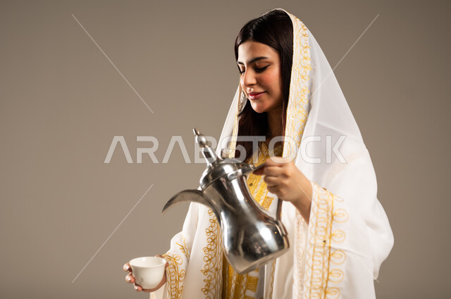 الأعياد و المناسبات الوطنية، مفهوم الكرم وحسن الاستقبال، يوم بدينا 1727 م، ذكرى تأسيس الدولة السعودية الاولى 22 فبراير، بورتريه مقرب لشابة عربية خليجية سعودية ترتدي العباءة البيضاء تقوم بصب القهوة من الدلة النحاسية، خلفية بيج