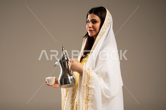 الأعياد و المناسبات الوطنية، مفهوم الكرم وحسن الاستقبال، يوم بدينا 1727 م، ذكرى تأسيس الدولة السعودية الاولى 22 فبراير، بورتريه مقرب لشابة عربية خليجية سعودية ترتدي العباءة البيضاء تقوم بصب القهوة من الدلة النحاسية، خلفية بيج