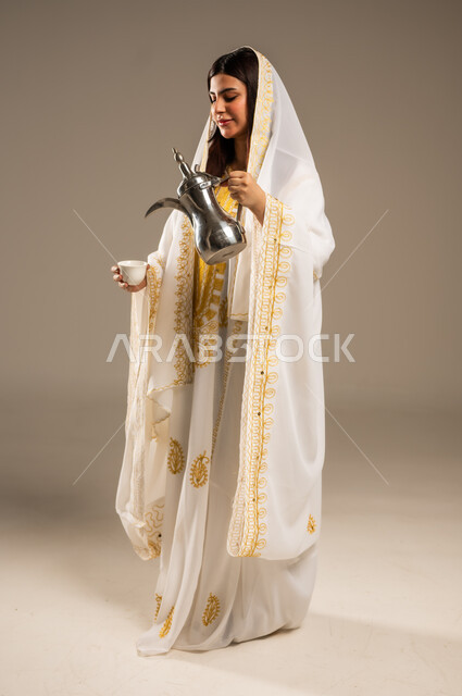 The concept of generosity and hospitality, the day we started 1727 AD, celebrating national occasions and holidays, the anniversary of the founding of the first Saudi state, February 22, a portrait of a young Saudi Gulf Arab woman wearing a white abaya pouring coffee from a copper pot, a picture of the body, beige background