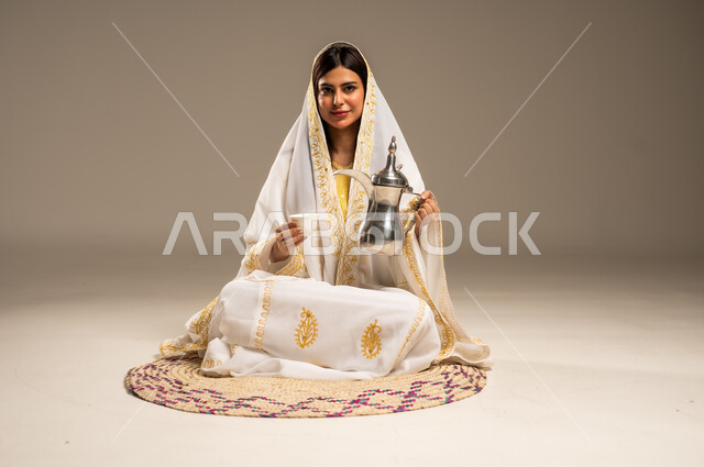 ذكرى تأسيس الدولة السعودية الاولى 22 فبراير، مفهوم الكرم وحسن الاستقبال، يوم بدينا 1727 م، بورتريه لشابة عربية خليجية سعودية ترتدي العباءة البيضاء تجلس على حصيرة من القش تحمل بيدها الدلة النحاسية تنظر الى الكاميرا بإيماءات السرور، خلفية بيج