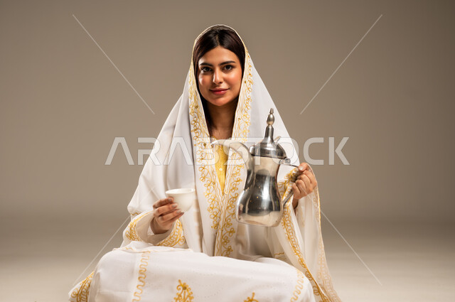 Anniversary of the founding of the first Saudi state, February 22, the concept of generosity and good reception, the day we started 1727 AD, celebrating national occasions and holidays, portrait of a young Saudi Gulf Arab woman wearing a white abaya holding a copper coffee pot in her hand looking at the camera with gestures of pleasure, beige background