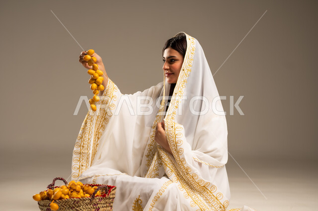 يوم بدينا 1727 م، المناسبات والأعياد الوطنية، منتجات ومحاصيل محلية، ذكرى تأسيس الدولة السعودية الاولى 22 فبراير، بورتريه لشابة عربية خليجية سعودية ترتدي العباءة البيضاء تحمل بيدها عنقود مليء بحبات البلح الطازجة، خلفية بيج