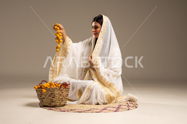 The Day We Began 1727 AD, National Events and Holidays, Local Products and Crops, Anniversary of the Founding of the First Saudi State February 22, Portrait of a young Saudi Arabian Gulf woman wearing a white abaya sitting on a straw mat holding a bunch of fresh dates in her hand, beige background