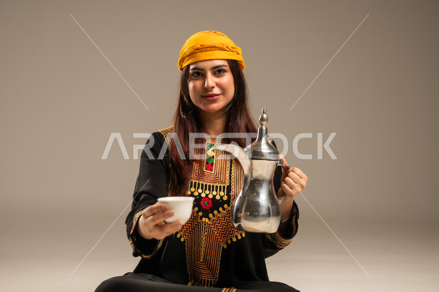 The concept of generosity and hospitality, love of the homeland and belonging to it, the anniversary of the founding of the first Saudi state, February 22, the day we started 1727 AD, a close-up portrait of a young Saudi Gulf Arab woman wearing a shayla and a sidr tree looking at the camera holding a copper coffee pot and a cup in her hand, beige background