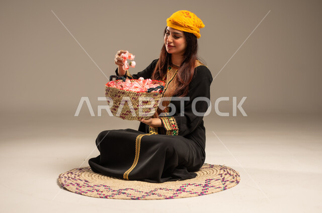 Anniversary of the founding of the first Saudi state, February 22, the concept of love of the homeland and belonging to it, national occasions and holidays, portrait of a young Saudi Gulf Arab woman wearing a shayla and a sidr tree sitting on a straw mat holding a basket full of natural roses, the day we began 1727 AD, beige background
