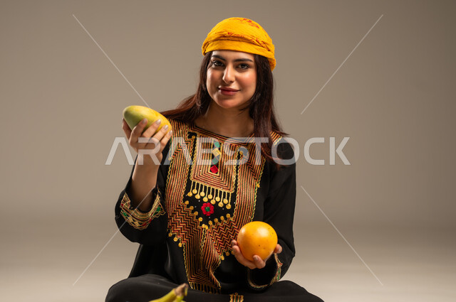The Day We Began 1727 AD, National Holidays and Occasions, Local Agricultural Crops and Products, Anniversary of the Founding of the First Saudi State February 22, Portrait of a Young Saudi Gulf Arab Woman Wearing a Shayla and a Sidra Tree Looking at the Camera Holding Fresh Fruits in Her Hand, Beige Background