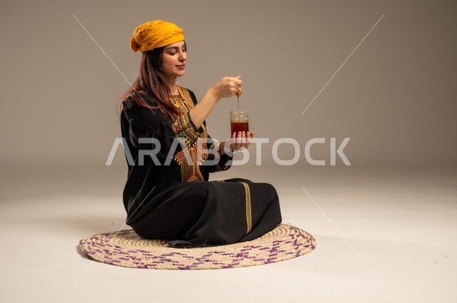 ذكرى تأسيس الدولة السعودية الاولى 22 فبراير، منتجات غذائية مفيدة، بورتريه لشابة عربية خليجية سعودية ترتدي الشيلة والسدرة تجلس على حصيرة من القش تحمل بيدها ملعقة مغمورة من عسل النحل الطبيعي في وعاء زجاجي، يوم بدينا 1727 م، المناسبات والأعياد الوطنية، خلفية بيج