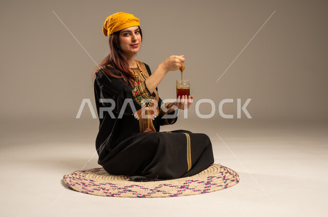 ذكرى تأسيس الدولة السعودية الاولى 22 فبراير، منتجات غذائية مفيدة، بورتريه لشابة عربية خليجية سعودية ترتدي الشيلة والسدرة تجلس على حصيرة من القش تنظر الى الكاميرا تحمل بيدها ملعقة مغمورة من عسل النحل الطبيعي في وعاء زجاجي، يوم بدينا 1727 م، خلفية بيج