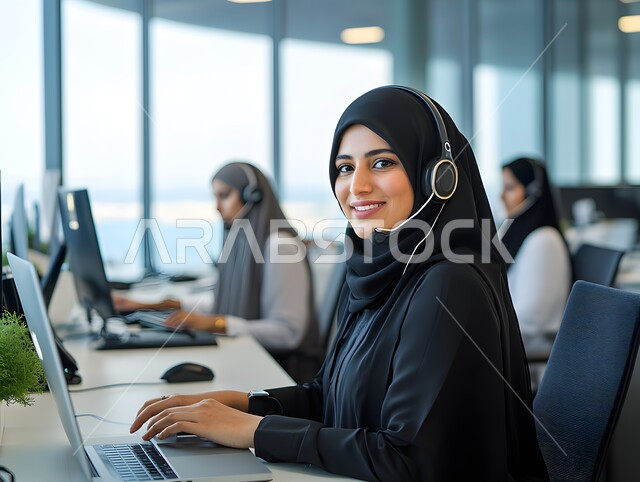 Answering and responding to inquiries, using advanced technologies in work, communicating with customers to solve problems, close-up of a smiling Saudi Arabian Gulf woman wearing a black abaya, wearing headphones, sitting inside the office and looking at the camera with gestures of happiness and pleasure