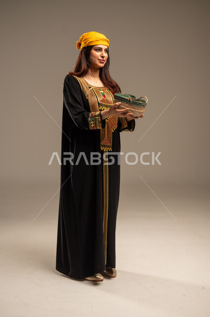 The Day We Started 1727 AD, Anniversary of the Founding of the First Saudi State February 22, Local Products and Crops, Celebrating National Occasions and Holidays, Portrait of a Young Saudi Arabian Gulf Woman Wearing a Shayla and a Sidra Tree Holding a Straw Basket Filled with Grains, Full-Length Body Image, Beige Background