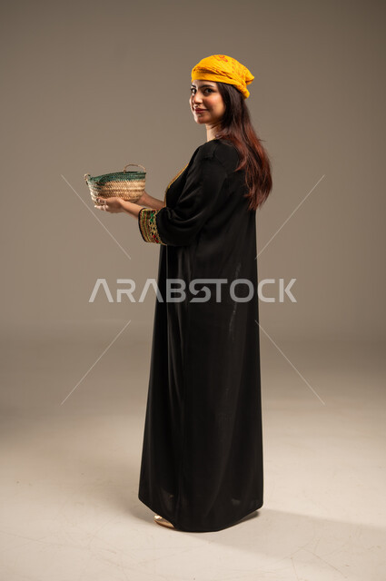 ذكرى تأسيس الدولة السعودية الاولى 22 فبراير، يوم بدينا 1727 م، منتجات ومحاصيل محلية، بورتريه من الجانب لشابة عربية خليجية سعودية ترتدي الشيلة والسدرة تنظر الى الكاميرا تحمل بيدها سلة من القش مليئة بالحبوب، صورة الجسم بالطول الكامل، خلفية بيج