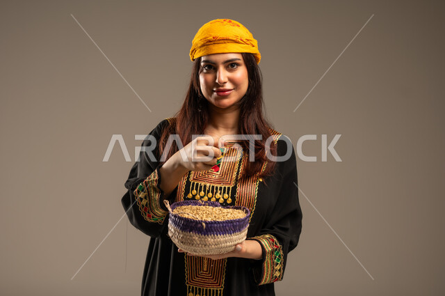 يوم بدينا 1727 م، منتجات ومحاصيل محلية، ذكرى تأسيس الدولة السعودية الاولى 22 فبراير، الاحتفال بالمناسبات الوطنية، بورتريه مقرب لشابة عربية خليجية سعودية ترتدي الشيلة والسدرة تنظر الى الكاميرا تحمل بيدها سلة من القش مليئة بالحبوب، خلفية بيج