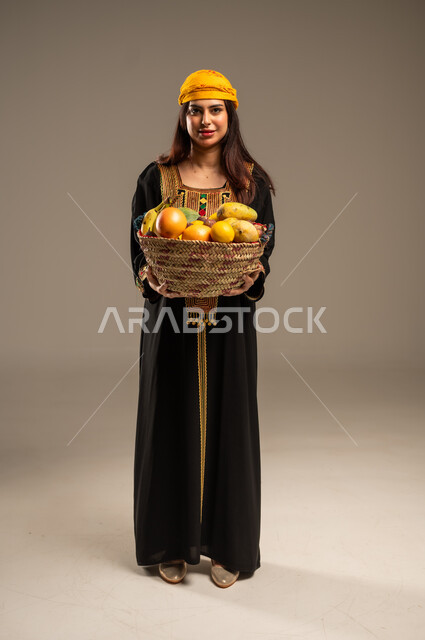 The Day We Began 1727 AD, Local Agricultural Crops and Products, Anniversary of the Founding of the First Saudi State February 22, Portrait of a young Saudi Arabian Gulf woman wearing a Shayla and Sidra tree looking at the camera holding a straw basket full of fresh fruits, full body image, beige background
