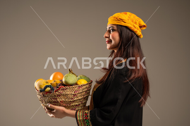 محاصيل و منتجات زراعية محلية، ذكرى تأسيس الدولة السعودية الاولى 22 فبراير، بورتريه مقرب من الجانب لشابة عربية خليجية سعودية ترتدي الشيلة والسدرة تحمل بيدها سلة من القش مليئة بالفواكه الطازجة، يوم بدينا 1727 م، خلفية بيج