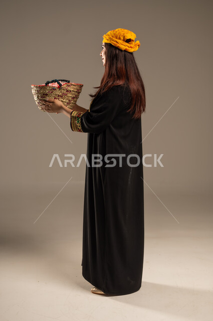 يوم بدينا 1727 م، ذكرى تأسيس الدولة السعودية الاولى 22 فبراير، بورتريه من الجانب لشابة عربية خليجية سعودية ترتدي الشيلة والسدرة تحمل بيدها سلة من القش ممتلئة بالورد الجوري الطبيعي، مفهوم حب الوطن والانتماء اليه، صورة الجسم بالطول الكامل، خلفية بيج