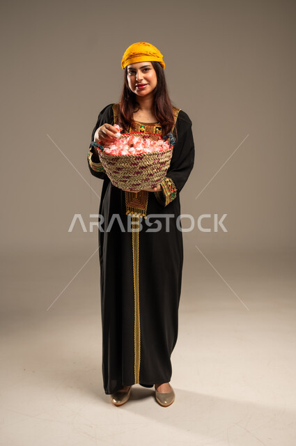 National occasions and holidays, the anniversary of the founding of the first Saudi state, February 22, a portrait of a young Saudi Gulf Arab woman wearing a shayla and a sidr tree looking at the camera holding a straw basket filled with natural roses, the day we began 1727 AD, a full-length body image, beige background