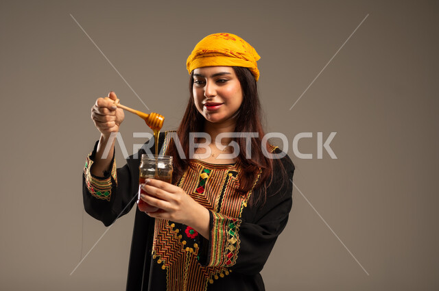 Anniversary of the founding of the first Saudi state February 22, the day we started 1727 AD, healthy and useful food products, celebrating national occasions, close-up portrait of a young Saudi Arabian Gulf woman wearing a shayla and a sidr tree holding a spoonful of natural honey in a glass jar, beige background