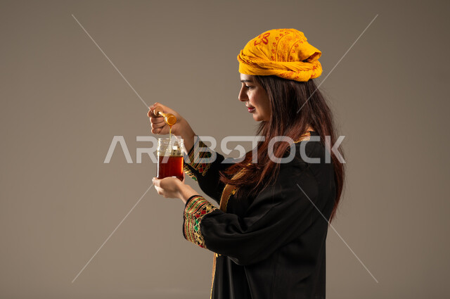 ذكرى تأسيس الدولة السعودية الاولى 22 فبراير، يوم بدينا 1727 م، منتجات غذائية صحية ومفيدة، الاحتفال بالمناسبات الوطنية، بورتريه مقرب من الجانب لشابة عربية خليجية سعودية ترتدي الشيلة والسدرة تحمل بيدها ملعقة مغمورة من عسل النحل الطبيعي في وعاء زجاجي، خلفية بيج