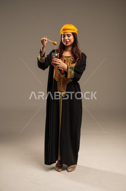 يوم بدينا 1727 م، ذكرى تأسيس الدولة السعودية الاولى 22 فبراير، منتجات غذائية صحية، الاحتفال بالمناسبات الوطنية، بورتريه لشابة عربية خليجية سعودية ترتدي الشيلة والسدرة تحمل بيدها ملعقة مغمورة من عسل النحل الطبيعي في وعاء زجاجي، صورة الجسم بالطول الكامل، خلفية بيج