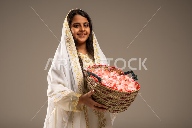 The concept of patriotism and belonging to the homeland, national holidays and occasions, the anniversary of the founding of the first Saudi state, February 22, a close-up portrait of a smiling Saudi Arabian Gulf girl wearing traditional costumes looking at the camera holding a basket of flowers in her hand, the day we began 1727 AD, beige background