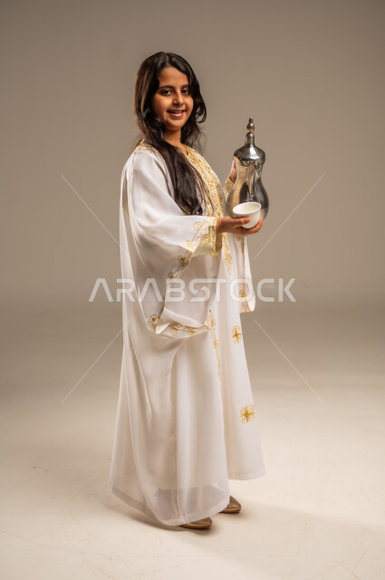 مفهوم الكرم وحسن الاستقبال، يوم بدينا 1727 م، الاحتفال بالمناسبات الوطنية، ذكرى تأسيس الدولة السعودية الاولى 22 فبراير، بورتريه لفتاة عربية خليجية سعودية ترتدي العباءة البيضاء تنظر الى الكاميرا تقوم بصب القهوة من الدلة النحاسية، صورة الجسم بالطول الكامل، خلفية بيج