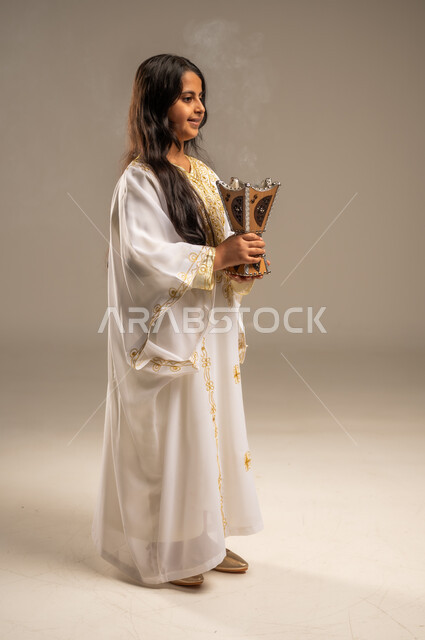The Day We Began 1727 AD, Using Agarwood to Perfume with Incense, Celebrating National Occasions and Holidays, Anniversary of the Founding of the First Saudi State February 22, Side Portrait of a Saudi Arabian Gulf Girl Wearing a White Abaya Holding an Incense Burner in Her Hand, Full-Length Body Image, Beige Background