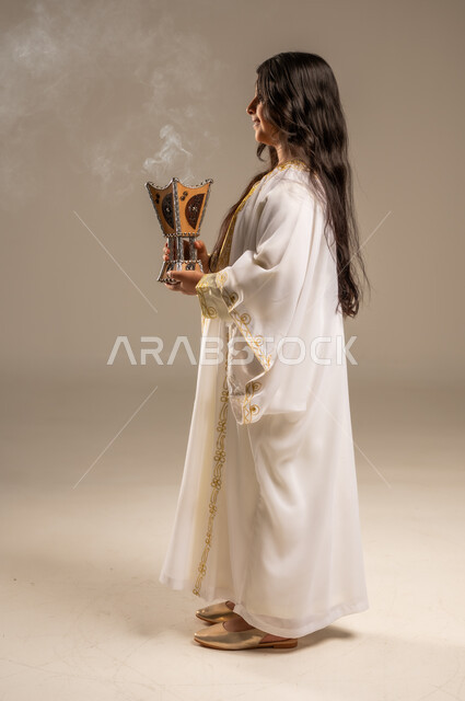 The Day We Began 1727 AD, Using Agarwood to Perfume with Incense, Celebrating National Occasions and Holidays, Anniversary of the Founding of the First Saudi State February 22, Side Portrait of a Saudi Arabian Gulf Girl Wearing a White Abaya Holding an Incense Burner in Her Hand, Full-Length Body Image, Beige Background