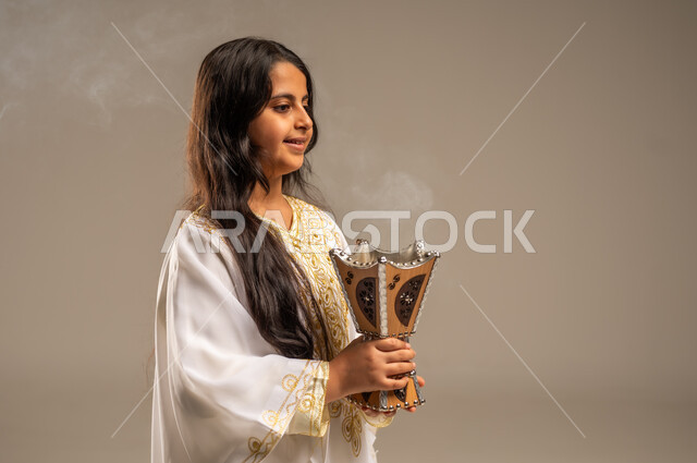 استخدام خشب العود للتطيب بالبخور، يوم بدينا 1727 م، الاحتفال بالمناسبات والأعياد الوطنية، ذكرى تأسيس الدولة السعودية الاولى 22 فبراير، بورتريه مقرب لفتاة عربية خليجية سعودية ترتدي العباءة البيضاء تحمل بيدها المبخرة، مفهوم حب الوطن والانتماء اليه، خلفية بيج