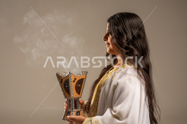 استخدام خشب العود للتطيب بالبخور، يوم بدينا 1727 م، الاحتفال بالمناسبات والأعياد الوطنية، ذكرى تأسيس الدولة السعودية الاولى 22 فبراير، بورتريه مقرب من الجانب لفتاة عربية خليجية سعودية ترتدي العباءة البيضاء تحمل بيدها المبخرة، مفهوم حب الوطن والانتماء اليه، خلفية بيج